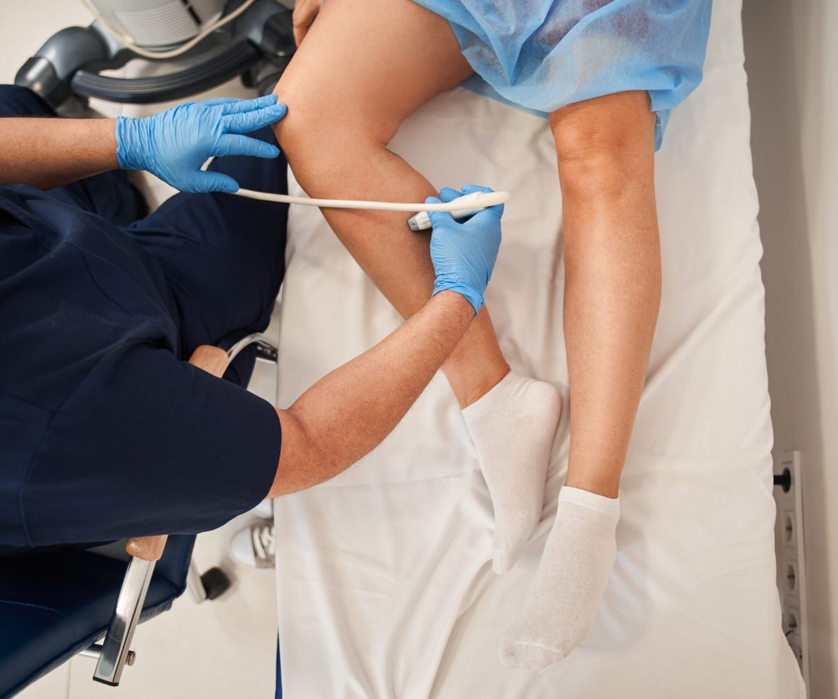 Orthopedist doctor doing ultrasound examination of patient’s leg veins in his office High angle view of the orthopedist doctor doing ultrasound examination of patient's leg veins in his office. Young woman passing ultrasound scan in clinic. Medical research concept